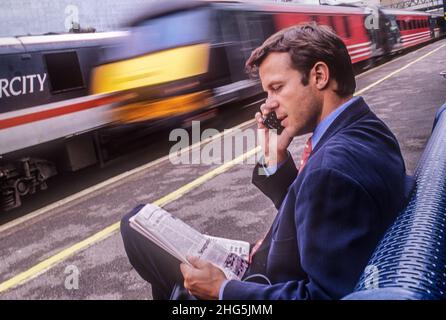 1990s Retro Travel homme d'affaires London train blur, parlant sur téléphone mobile assis sur le banc de plate-forme de la gare britannique. Lire un journal et parler de la dernière technologie 90s petit téléphone mobile Ericsson. Le train de la Vierge inter-ville de Cross Country passe avec un flou de vitesse. La technologie de mode Voyage et transport dans le 1990s Banque D'Images