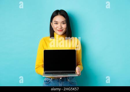 Belle et élégante femme asiatique montrer le produit à l'écran, montrant l'écran vide d'ordinateur portable et souriant, debout sur fond bleu Banque D'Images