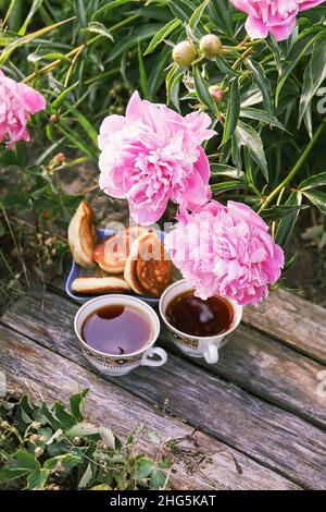 Thé de style campagnard dans le jardin d'été du village.Deux tasses de thé noir chaud et des crêpes sur des planches de bois abîmés et fleur de pivoine rose en fleur Banque D'Images