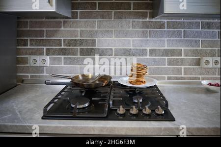 Vue de face de crêpes maison fraîchement cuites sur plaque blanche, poêle en acier et spatule sur une cuisinière noire dans la cuisine.Le mardi de Shrove concep Banque D'Images