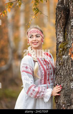 Jeune belle femme souriante en folk traditionnelle robe slave embrasse l'arbre dans la nature en forêt d'automne.Portrait d'une jolie fille en vêtements ethniques Banque D'Images