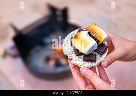 Jeune femme campant de près tenant main des marshmallows rôtis avec des carrés de barre de chocolat et des craquelins de gâteau de riz par une fosse de feu dans le camping Banque D'Images