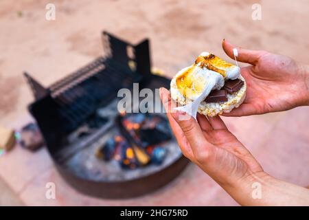 Femme gros plan main tenant des marshmallows collants rôtis avec des taches de riz au chocolat papillote de gâteau par foyer dans le camping-grill feu Banque D'Images