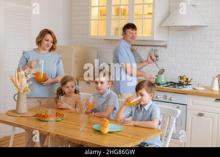 Bonne famille caucasienne avec des enfants assis à la table de cuisine et de boire du jus frais ensemble, maman souriante s'amusant avec sa fille et ses fils, appréciant un week-end familial à la maison Banque D'Images