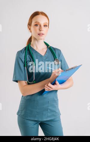 Portrait vertical de la jeune femme médecin sérieux en uniforme médical écrivant la prescription sur le presse-papiers, remplit l'histoire médicale, regardant la caméra Banque D'Images