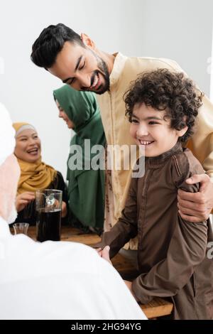 Homme arabe souriant qui embrasse son fils près du thé et de la famille multiethnique à la maison Banque D'Images