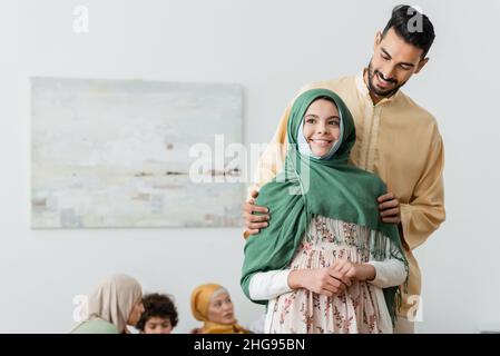 un homme arabe heureux embrassant une fille musulmane près d'une famille multiethnique floue à la maison Banque D'Images