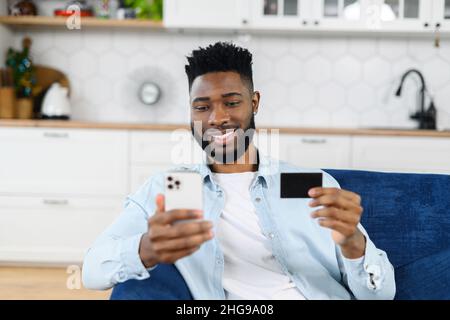 Souriant homme multiracial shopping en ligne assis sur le canapé à la maison, tenant la carte de crédit et le smartphone.Un gars multiracial à la peau sombre qui commande des aliments, achète en ligne, effectue des réservations et paie Banque D'Images