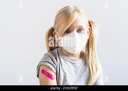 petite fille blonde portant un masque de visage montrant son bras avec bandage rose après avoir été vaccinée ou inoculation, vaccination des enfants, covid delta vaccin con Banque D'Images