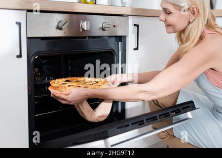 Vue latérale d'une femme mûre positive prenant une tarte du four dans la cuisine Banque D'Images