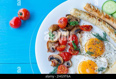 Petit déjeuner composé d'œufs frits, saucisses grillées, champignons frits et tomates cerises, tranches de concombre fraîches sur une assiette blanche sur une table en bois bleu.Voir fr Banque D'Images