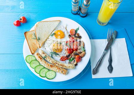 Petit-déjeuner composé d'œufs frits, saucisses grillées, champignons frits et tomates cerises, tranches de concombre frais et pain grillé sur une assiette blanche sur une bl en bois Banque D'Images