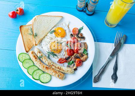 Petit-déjeuner composé d'œufs frits, saucisses grillées, champignons frits et tomates cerises, tranches de concombre frais et pain grillé sur une assiette blanche sur une bl en bois Banque D'Images