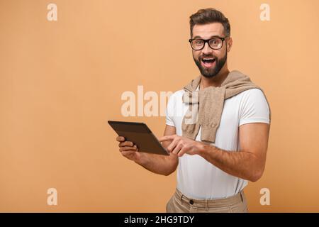 Portrait de l'homme barbu débordé avec tablette se réjouissant de quelque chose en posant isolé sur fond orange Banque D'Images