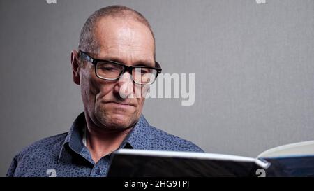 Homme senior dans des lunettes élégantes lit le livre sur fond gris Banque D'Images