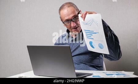 Un homme âgé vérifie les dessins près d'un ordinateur portable au bureau blanc Banque D'Images