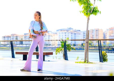 Portrait de la belle fille blonde adolescente avec des queues de porc fraîches, t-shirt blanc et pantalon évasé rose patinage sur roulettes dans le parc Banque D'Images