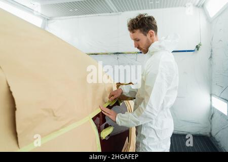 Mécanicien de sexe masculin couvrant le véhicule de carrosserie avec du papier et du ruban de masquage avant de peindre en service de réparation automobile Banque D'Images