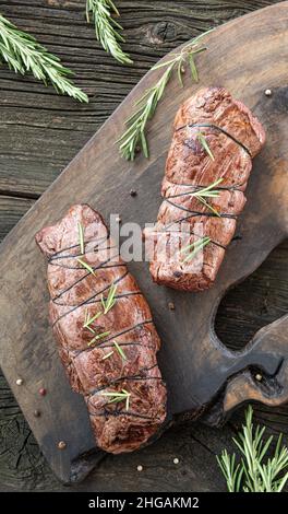 Deux portions de filets de bœuf fraîchement rôtis juteux, nouées avec du fil de cuisson, du poivre noir et des tiges de romarin vert frais sur du cuttin en bois Banque D'Images