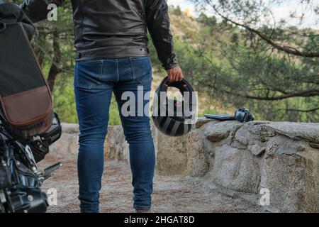 Un motocycliste en Jean avec un casque dans les mains se trouve à la moto dans le parking. Banque D'Images