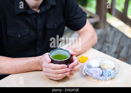 Homme assis à la table, près des mains tenant une tasse de thé vert matcha à l'extérieur dans le jardin de l'arrière-cour avec la nourriture japonaise daifuku mochi Banque D'Images