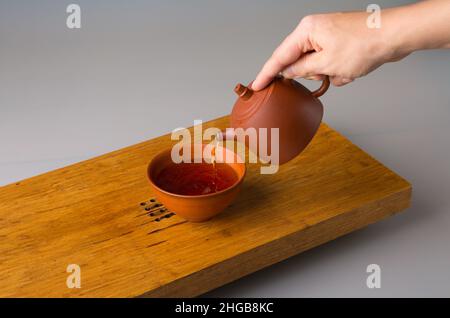 La main d'un homme verse du thé pu erh fraîchement préparé d'une théière en argile dans une tasse traditionnelle.Une tasse sur une table pour les cérémonies du thé. Banque D'Images