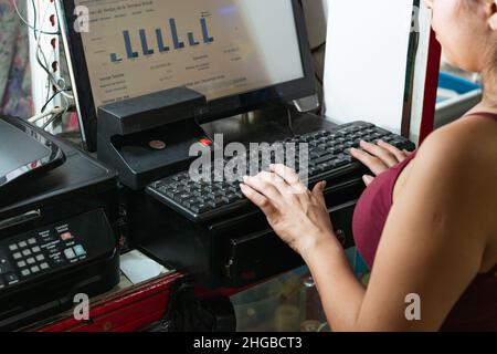 latina femme entrepreneur dans son entreprise regardant le graphique des statistiques de ventes sur l'ordinateur. fille devant la caisse de trésorerie à l'aide de son clavier Banque D'Images