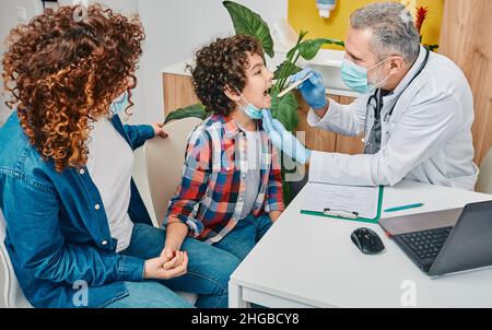 Enfant avec son parent au rendez-vous pédiatre.Un généraliste, utilisant une spatule d'inspection, examine la gorge du petit garçon bouclés Banque D'Images