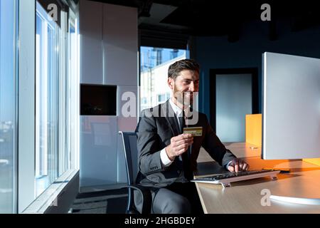 joyeux homme d'affaires barbu shopping en ligne travaillant dans un ordinateur de bureau moderne, homme tenant une carte de crédit bancaire Banque D'Images