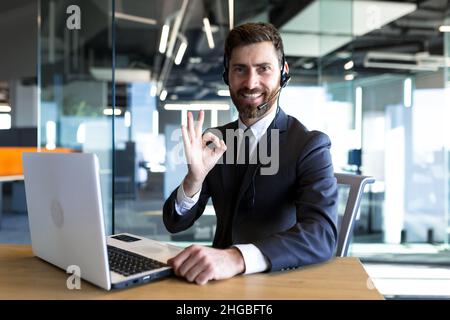 Un employé de centre d'appels heureux et réussi regarde la caméra et sourit, l'homme d'affaires garde son doigt à portée de main, travaillant sur l'ordinateur, utilise un Banque D'Images