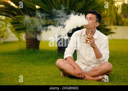 Un homme de vape en vacances, sous un palmier.Repos, tabagisme, nicotine, plaisir, danger.Liquide VAPE.Vaping THC e cigarette. Banque D'Images