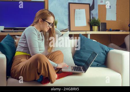 Une fille travaille à la maison, est assise sur un canapé avec un ordinateur.École à domicile, école en ligne.Une fille regarde un ordinateur, fait des achats sur Internet.Restez h Banque D'Images