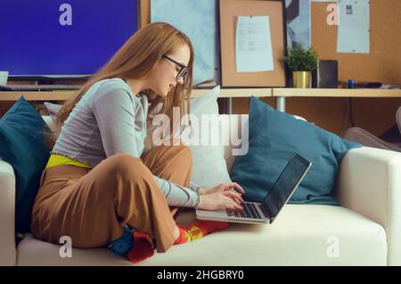 Une fille travaille à la maison, est assise sur un canapé avec un ordinateur.École à domicile, école en ligne.Une fille regarde un ordinateur, fait des achats sur Internet.Restez h Banque D'Images