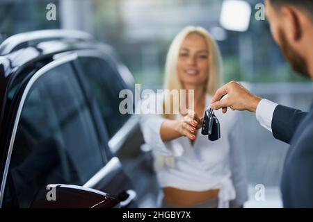 Joyeuse femme caucasienne prenant les clés de sa nouvelle voiture tout en étant debout avec un consultant masculin dans un salon moderne.Concept de personnes, achat et vente. Banque D'Images