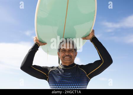 Portrait d'un homme biracial senior souriant portant une planche de surf sur la tête à la plage ensoleillée contre le ciel Banque D'Images