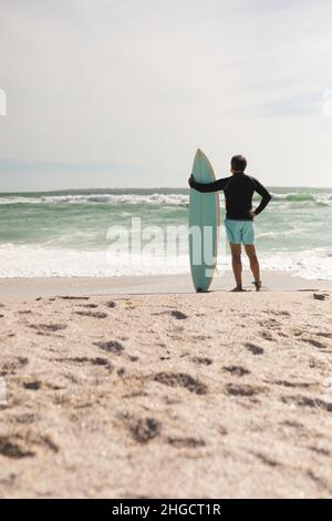 Vue arrière de l'homme biracial senior debout avec planche de surf sur le rivage à la plage pendant la journée ensoleillée Banque D'Images