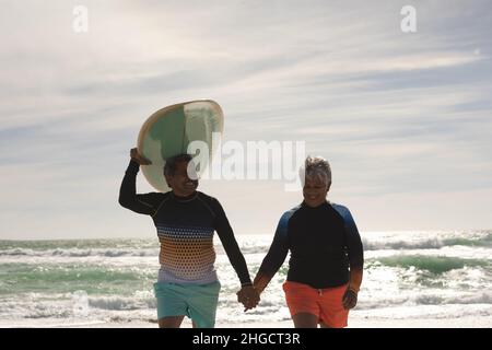 Homme biracial senior souriant portant une planche de surf sur la tête tout en tenant la main de la femme marchant à la plage Banque D'Images