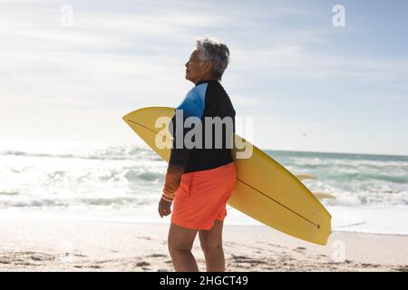 Vue latérale d'une femme âgée biraciale portant une planche de surf et regardant la plage pendant la journée ensoleillée Banque D'Images