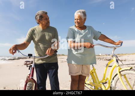 Couple multiracial senior regardant l'un l'autre debout avec des vélos sur la plage ensoleillée contre le ciel Banque D'Images