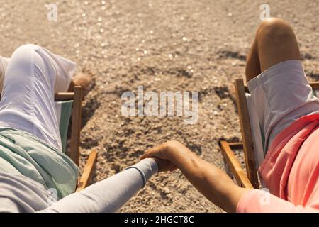 Vue en grand angle d'un couple multiracial senior tenant les mains tout en se relaxant sur des chaises à la plage Banque D'Images