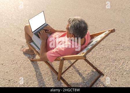 Vue en grand angle de l'homme aîné biracial à l'aide d'un ordinateur portable à écran vierge avec espace pour copier sur une chaise à la plage Banque D'Images