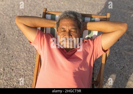 Vue en grand angle de l'homme biracial senior dormant avec les mains derrière la tête sur une chaise se relaxant à la plage Banque D'Images