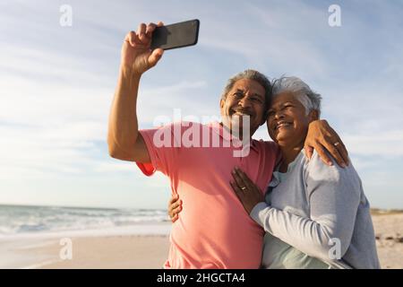 Homme biracial senior souriant prenant le selfie à travers le smartphone avec le bras autour de la femme à la plage Banque D'Images
