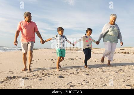 Pleine longueur d'homme et de femme biracial senior tenant les mains avec des petits-enfants tout en marchant à la plage Banque D'Images