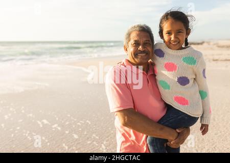 Portrait d'un homme biracial senior souriant portant la petite-fille à la plage contre le ciel Banque D'Images