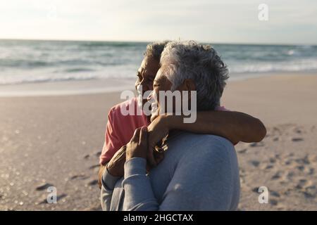 Couple biracial senior souriant s'embrassant tout en regardant loin debout ensemble à la plage Banque D'Images