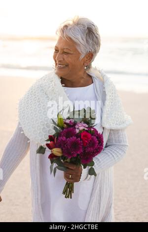 Femme biraciale senior souriante avec cheveux blancs tenant un bouquet de fleurs donnant sur la plage Banque D'Images