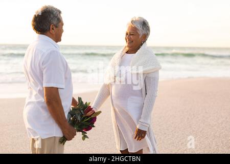 Femme biraciale senior souriante tenant la main de l'homme avec un bouquet à la plage pendant la cérémonie de mariage Banque D'Images