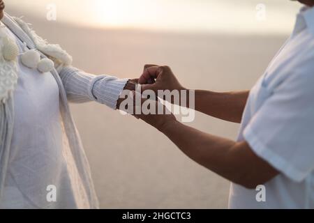 Section médiane de la groom biracial senior mettant l'anneau de mariage sur le doigt de la mariée à la plage pendant le coucher du soleil Banque D'Images