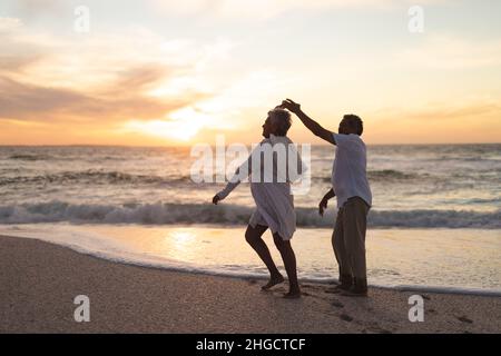 Couple multiracial senior heureux dansant sur la plage contre le ciel pendant le coucher du soleil Banque D'Images
